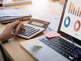 hand using calculator next to laptop displaying budget graphs